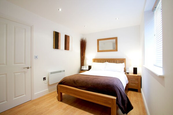 A bedroom with a wooden bed, white walls, a radiator, a nightstand, a mirror, and a window with blinds.