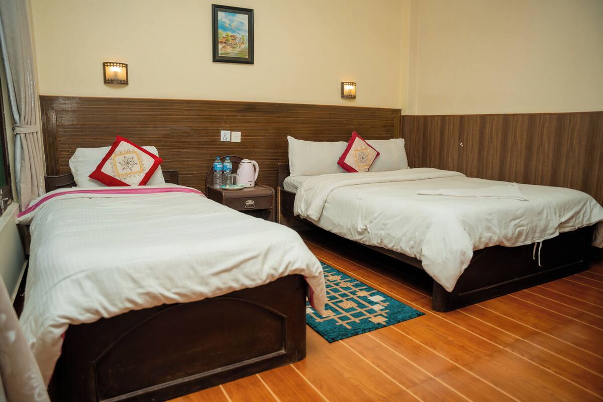A hotel room with two beds, wooden headboard, bedside tables, a nightstand, a blue rug, and wall-mounted lights.
