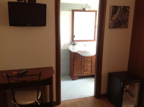 Une pièce avec un bureau en bois, un téléviseur fixé au mur et une salle de bain visible à travers une porte ouverte.