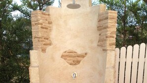 Une douche extérieure en pierre, avec un sol carrelé, entourée de verdure et d’une clôture en bois peinte en blanc.