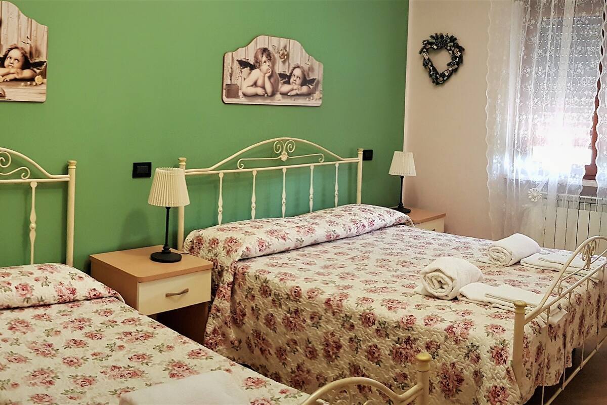 Ein Schlafzimmer mit zwei Einzelbetten, einem Nachttisch mit Lampe, floralem Bettbezug und Wanddekorationen, darunter Darstellungen von Engeln.