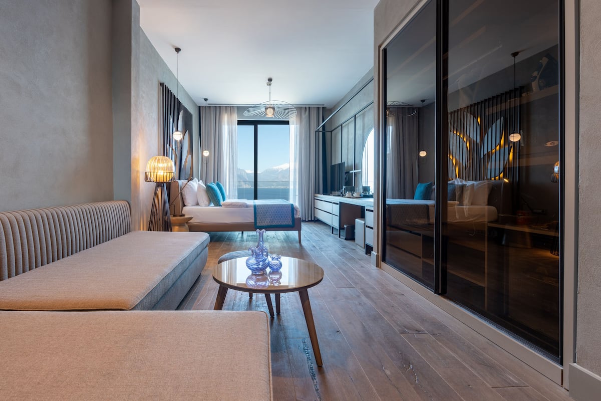 A modern hotel room with a sofa, a small table, a view of the mountains, and a large window.