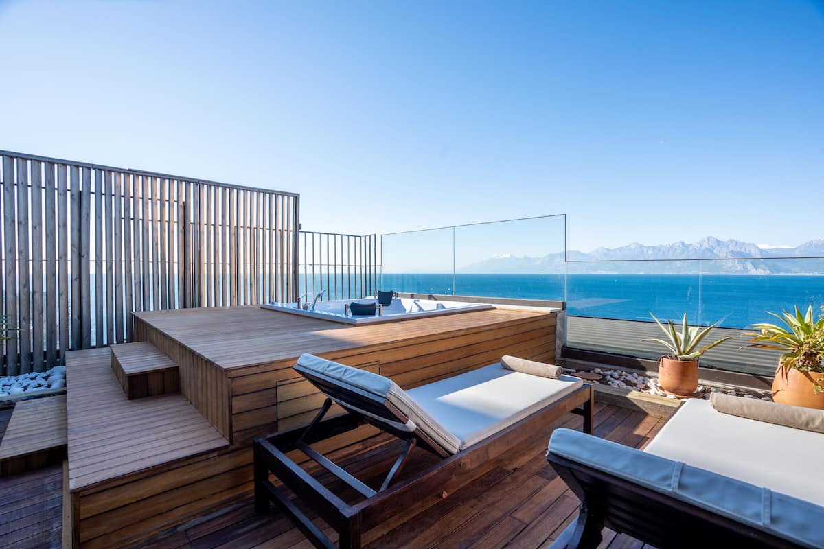 A wooden deck with lounge chairs and a small seating area overlooking a body of water with mountains in the background.