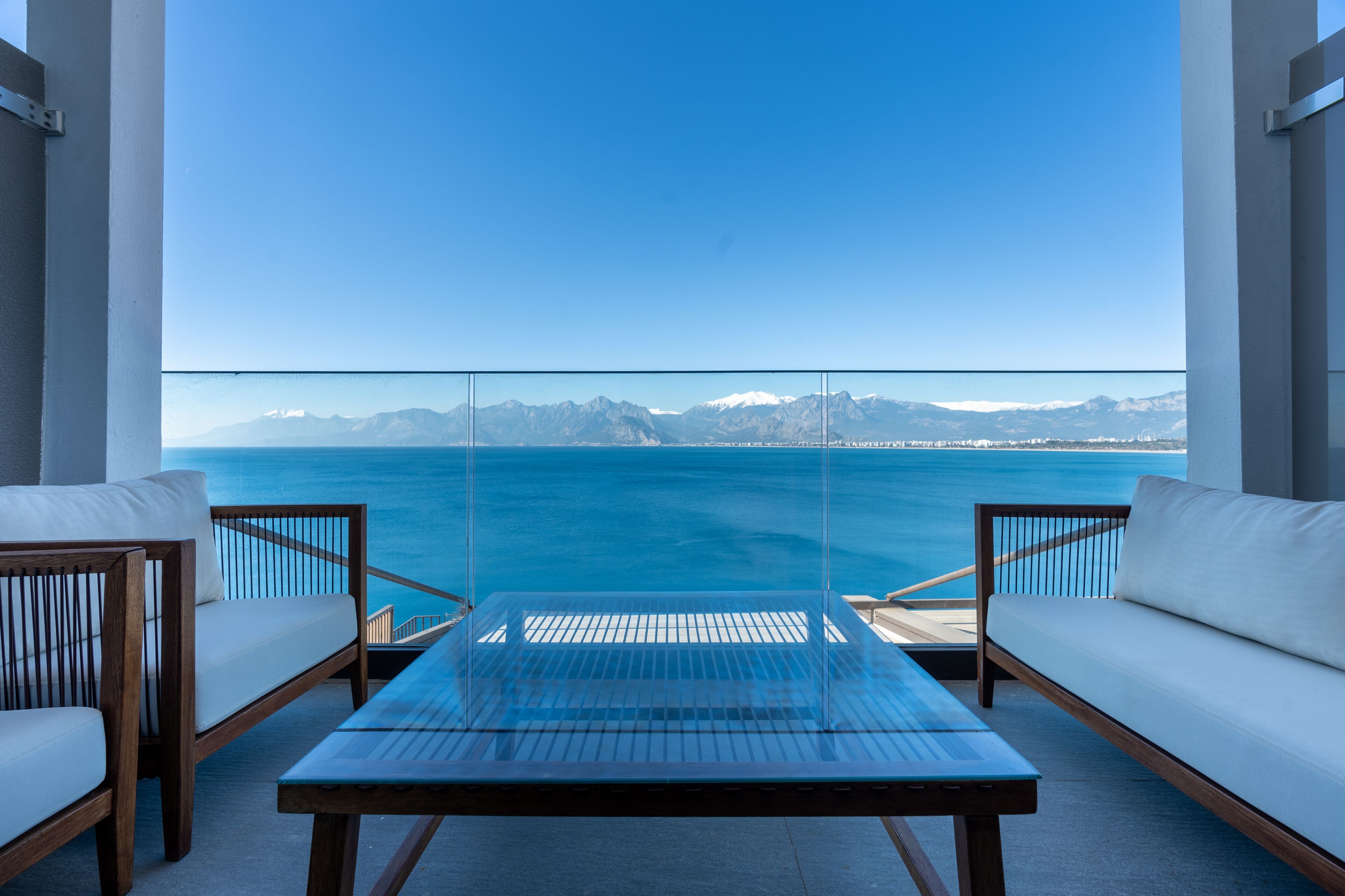 A balcony with a view of the sea and mountains, featuring a glass table and two white sofas.
