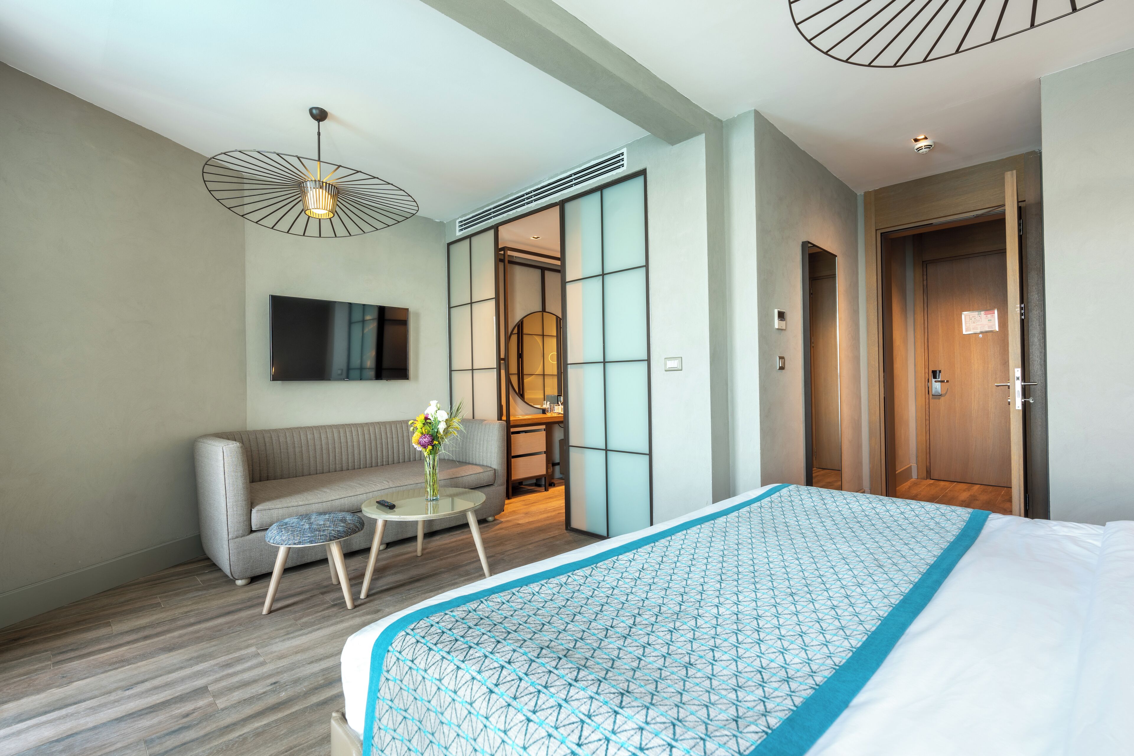 A modern hotel room with a large bed, a sofa, a small table with a vase of flowers, a TV mounted on the wall, and a sliding door leading to a bathroom.