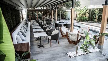 A covered outdoor seating area with tables and chairs, surrounded by plants and a view of a pool.