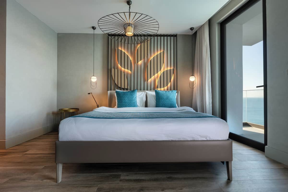 A modern bedroom with a large bed, a ceiling light fixture, and a view of the ocean through a large window.