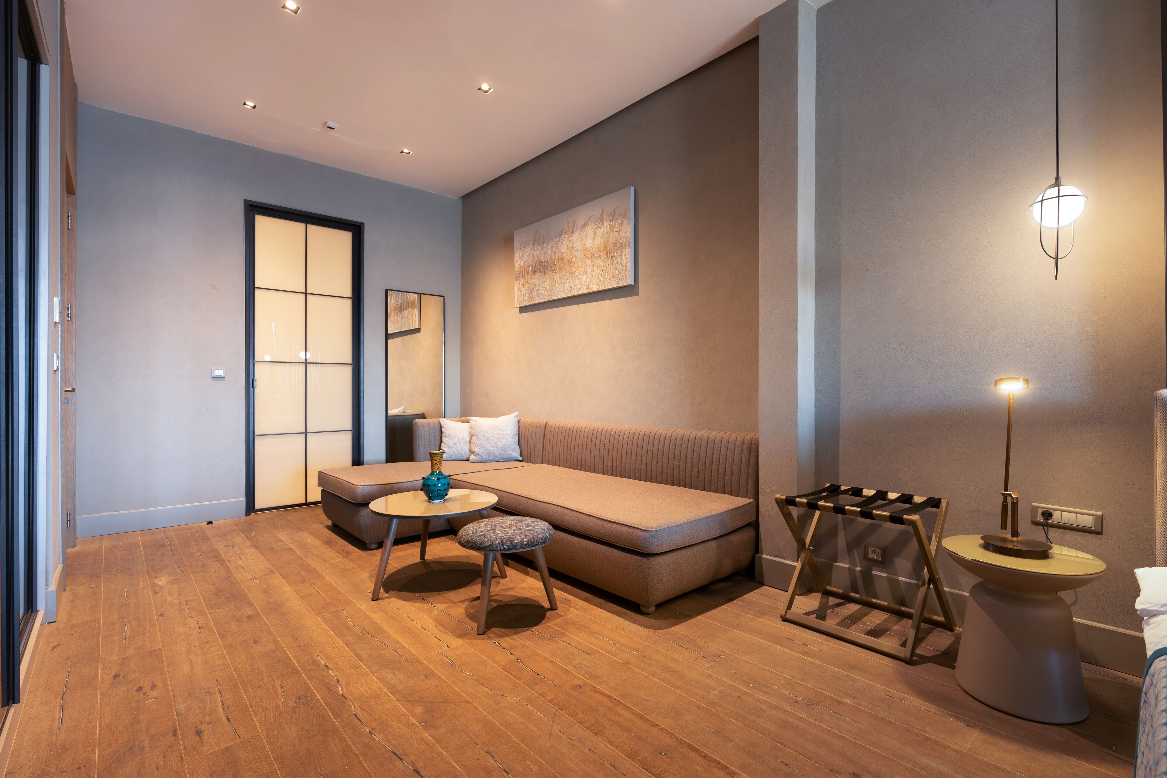 A modern living room with a sofa, a round table, and a small stool. There is a wall-mounted artwork and a floor lamp.