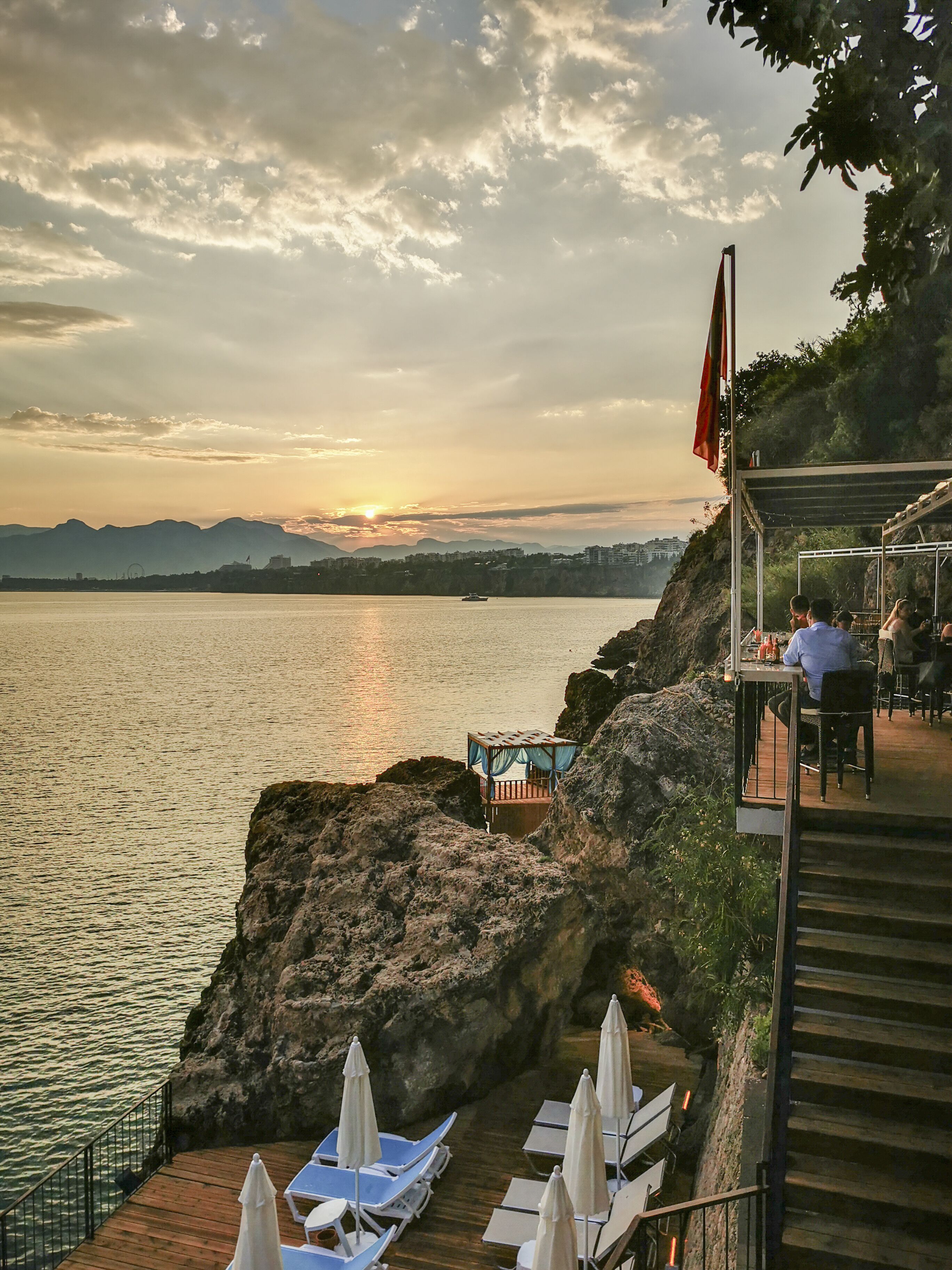 Una terraza frente al mar con mobiliario y vista al atardecer sobre el agua.