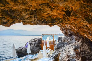 A beach with lounge chairs and umbrellas under a rock formation. - Perge Hotels Adult Only (Antalya)