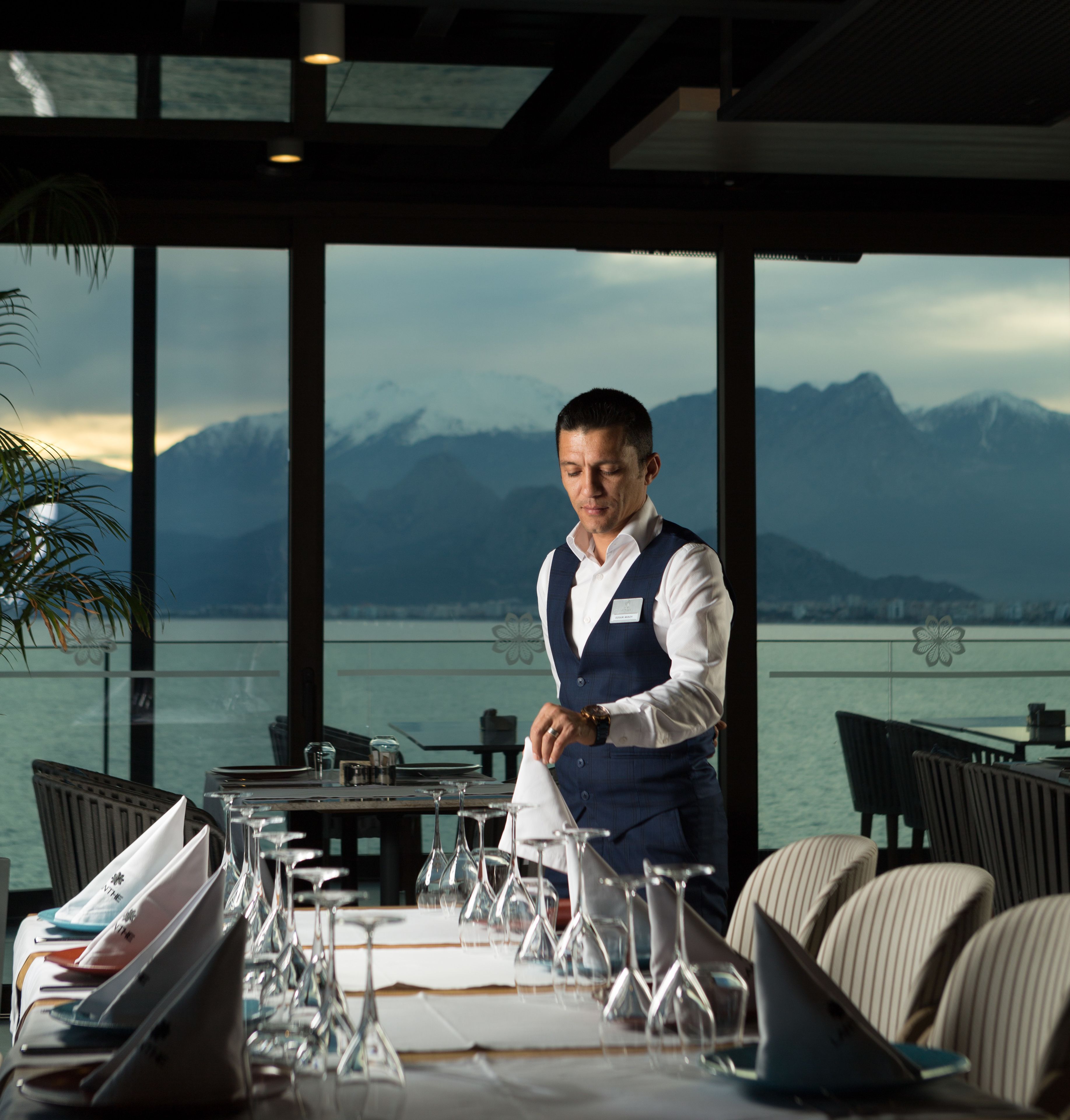 Un camarero prepara una mesa con copas de vino y un menú en un restaurante con vistas a las montañas y al agua.