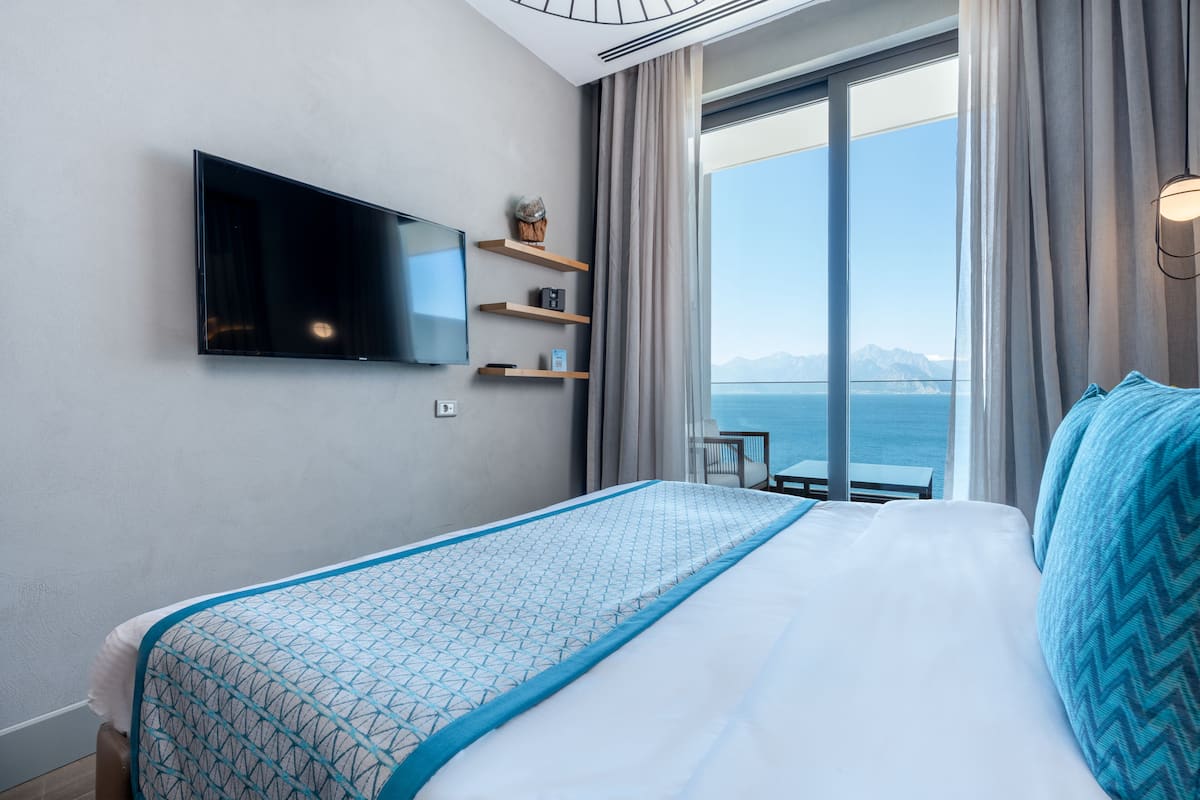 A hotel room with a bed, a TV mounted on the wall, a small table, and a view of the sea through large windows.