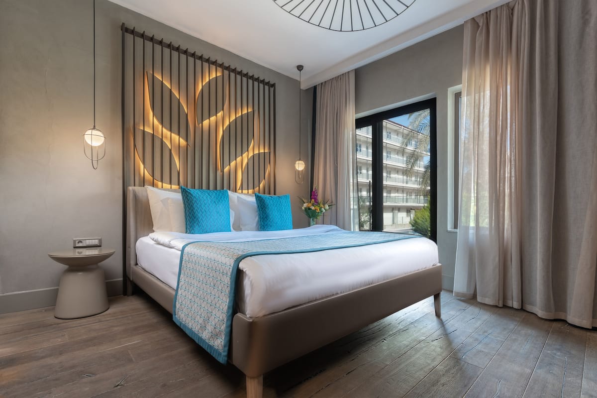 A modern hotel room with a large bed, a bedside table, a wall-mounted light, and a view of the outside.