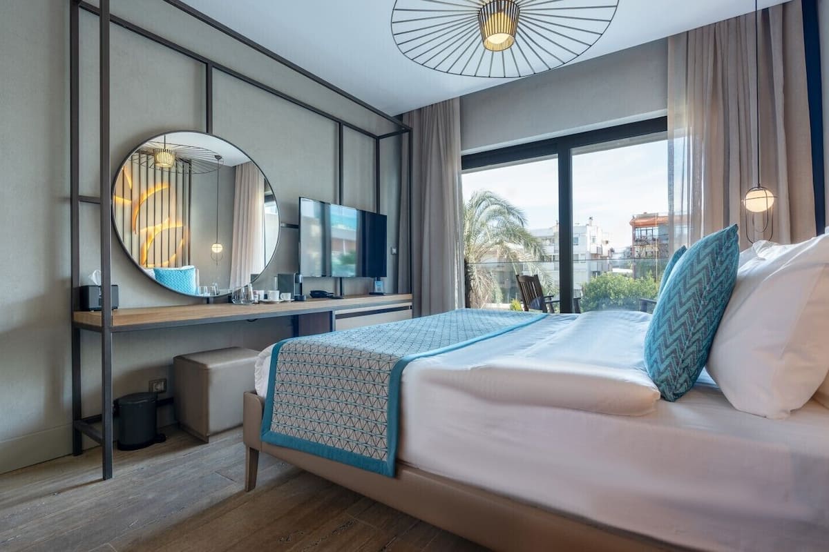 A modern hotel room with a large bed, a desk with a flat-screen TV, a round mirror, and a view of the city.