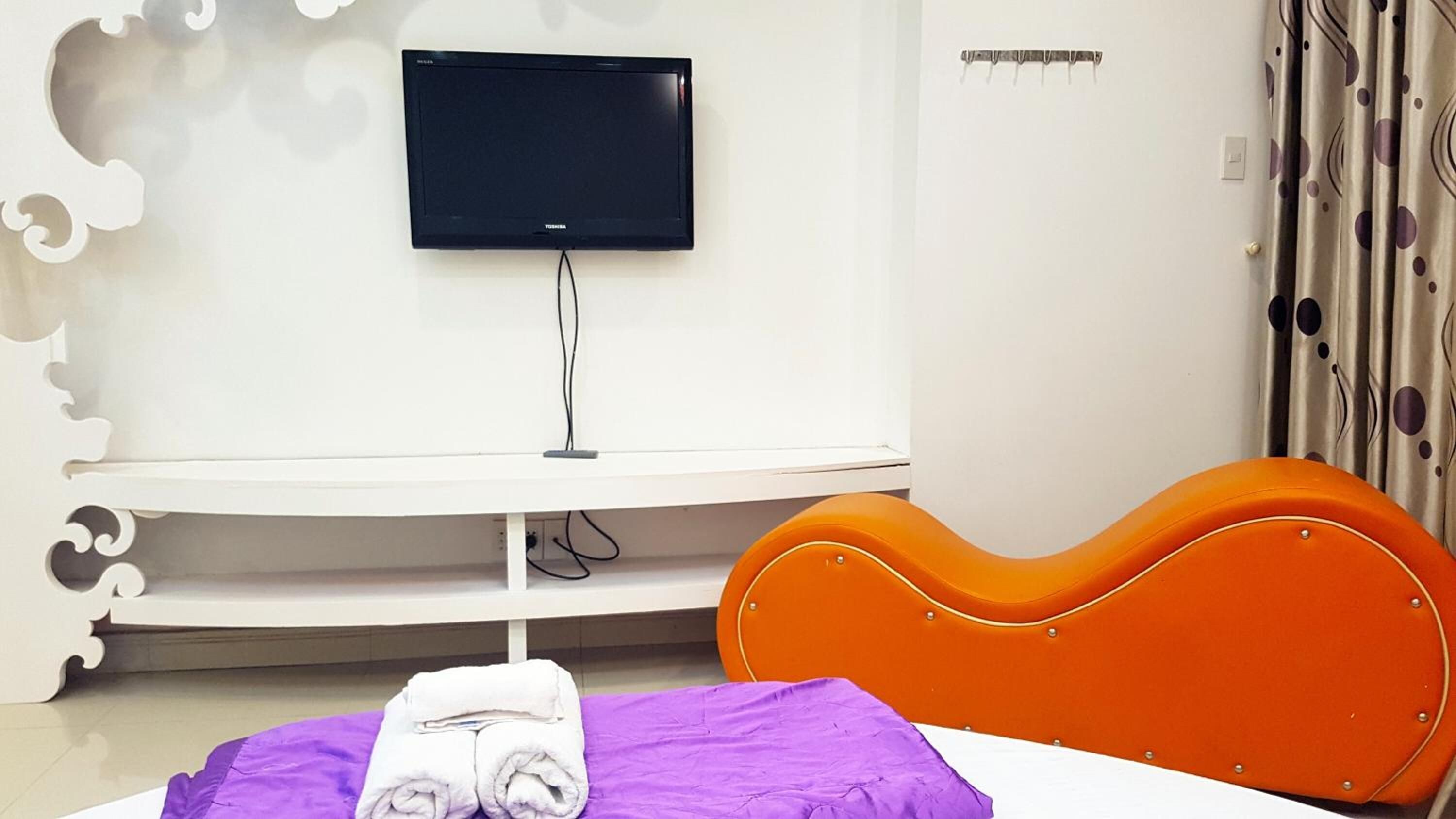 A hotel room with a flat-screen TV mounted on a white wall, a white console table, an orange headboard, and a purple bedspread with rolled towels.
