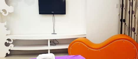 A hotel room with a flat-screen TV mounted on a white wall, a white console table, an orange headboard, and a purple bedspread with rolled towels.
