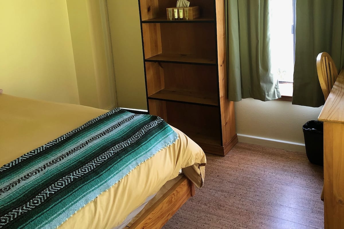 Ein Zimmer mit einem Bett, einem Holzregal, einem Fenster mit Vorhängen und einem kleinen Tisch mit Stuhl.