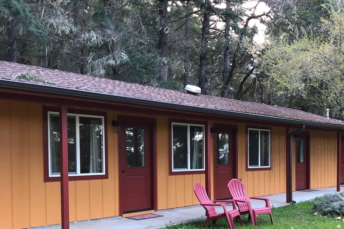 Ein eingeschossiges Gebäude mit roter Tür und Fenstern, einer kleinen Veranda mit zwei pinkfarbenen Adirondack-Stühlen und einem Rasen.