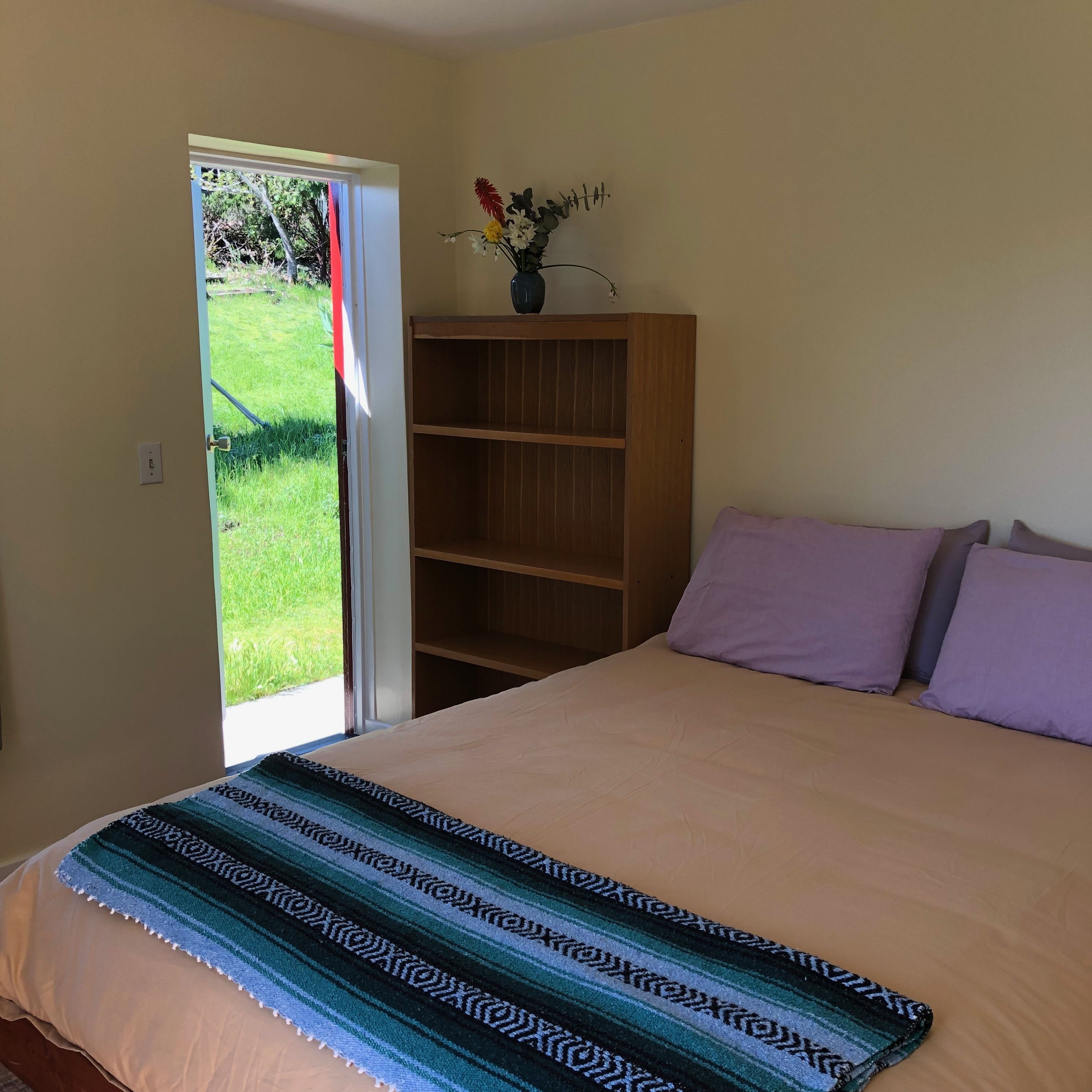 A bedroom with a bed, a wooden bookshelf, and a window with a view of a garden.