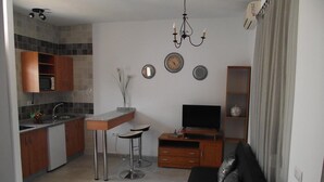 A modern kitchen with a microwave, sink, and bar stools. There is a dining table with chairs, a TV on a wooden stand, and a black sofa with decorative pillows. - Hostal El Canario (Conil de la Frontera)