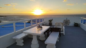 En terrasse med bord og stole med udsigt over havet i solnedgang.