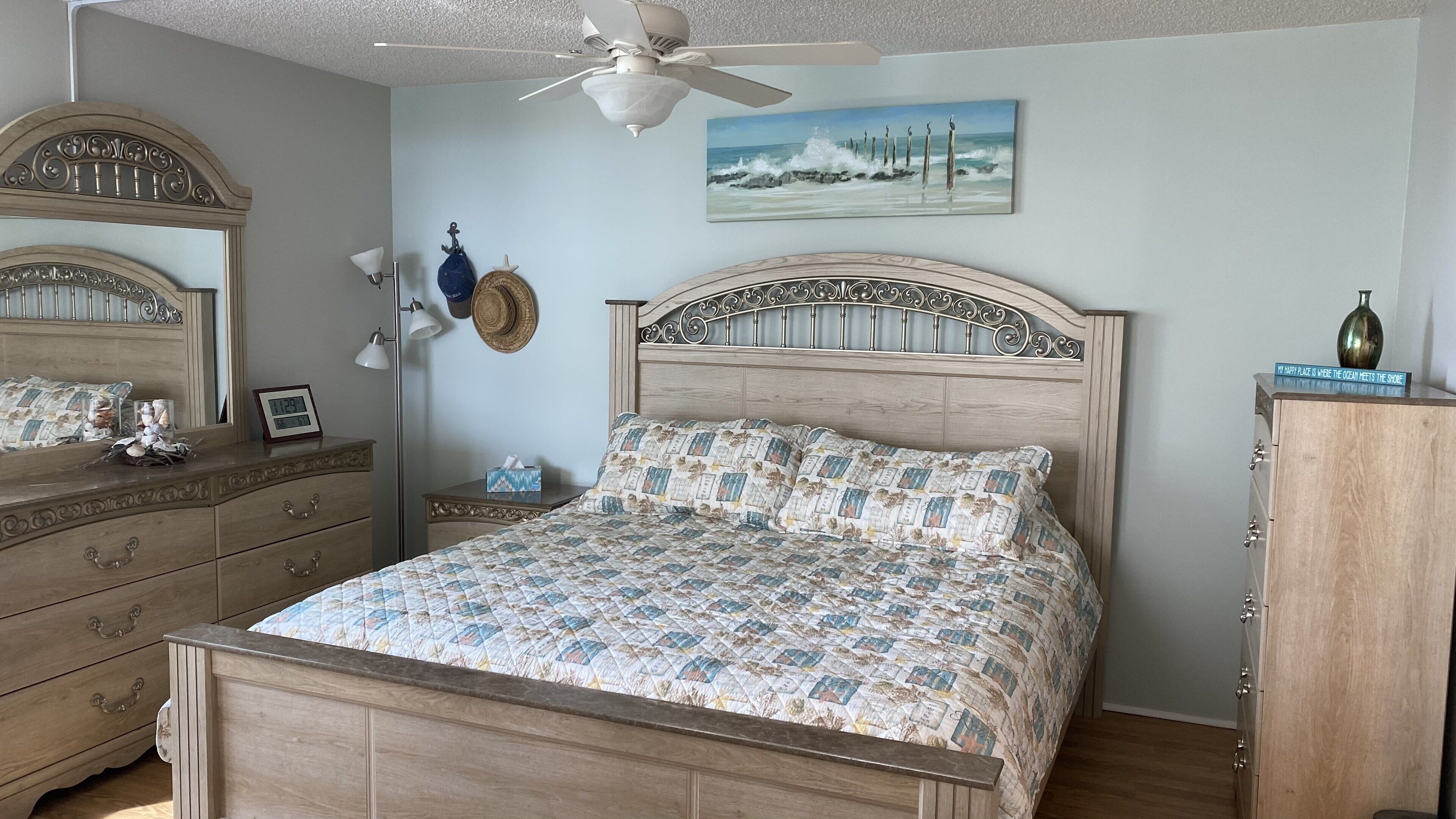 Un dormitorio con una cama de madera, una mesita de noche, un aparador, un espejo, un ventilador de techo y un cuadro de una escena de playa.