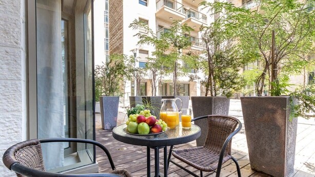 A balcony with wicker chairs, a table with fruit and juice, and a view of a building with balconies and trees.