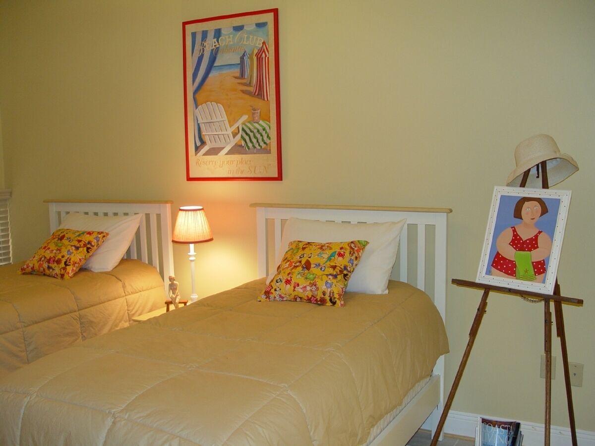 Une chambre d’hôtel avec deux lits, un tableau sur le thème de la plage, un petit chevalet sur lequel est posé un portrait, et une lampe.