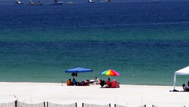 Ein Strand mit mehreren Booten im Wasser, Menschen entspannen unter Sonnenschirmen, und ein sandiger Uferstreifen.