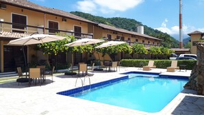A pool area with lounge chairs and umbrellas, surrounded by a building with a tiled roof and a mountain in the background. - Charming house in a gated community in Juquehy. - 2 suites (São Sebastião)