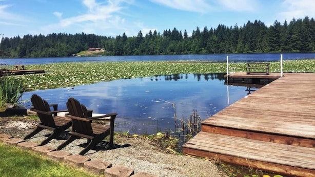 A wooden deck with chairs and a small table, overlooking a lake with lily pads and a forested shoreline. - Lakeside Living Suite! ~Mt. Rainier, NWTrek, JBLM (Graham)