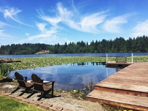 Una terraza de madera con sillas y una mesita pequeña, con vistas a un lago cubierto de lirios y una orilla boscosa.