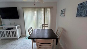 A dining area with a dark wooden table, white chairs, a ceiling fan, a TV on a white stand, and a window with blinds.