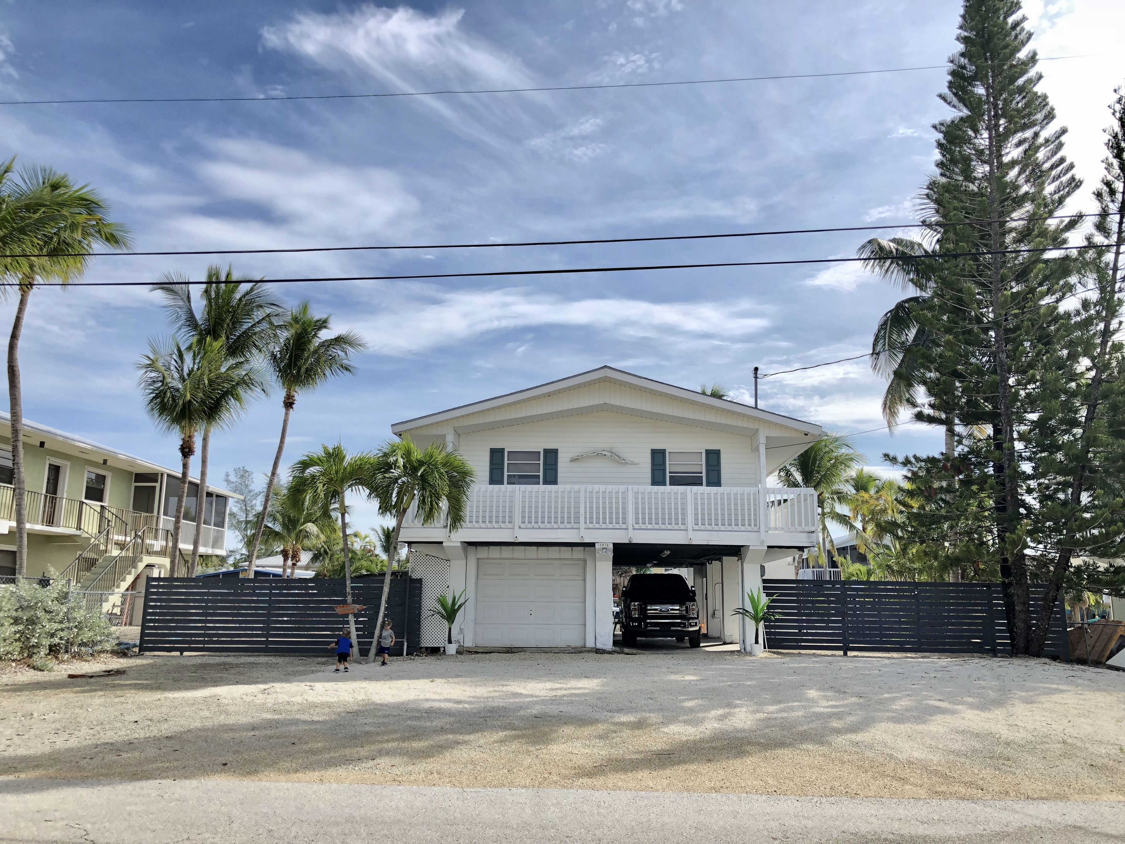 A two-story house with a garage, surrounded by palm trees and a fence.