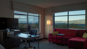 A hotel room with a desk, chair, red sofa, and a view of mountains through large windows. - Snoqualmie Inn by Hotel America (Snoqualmie)