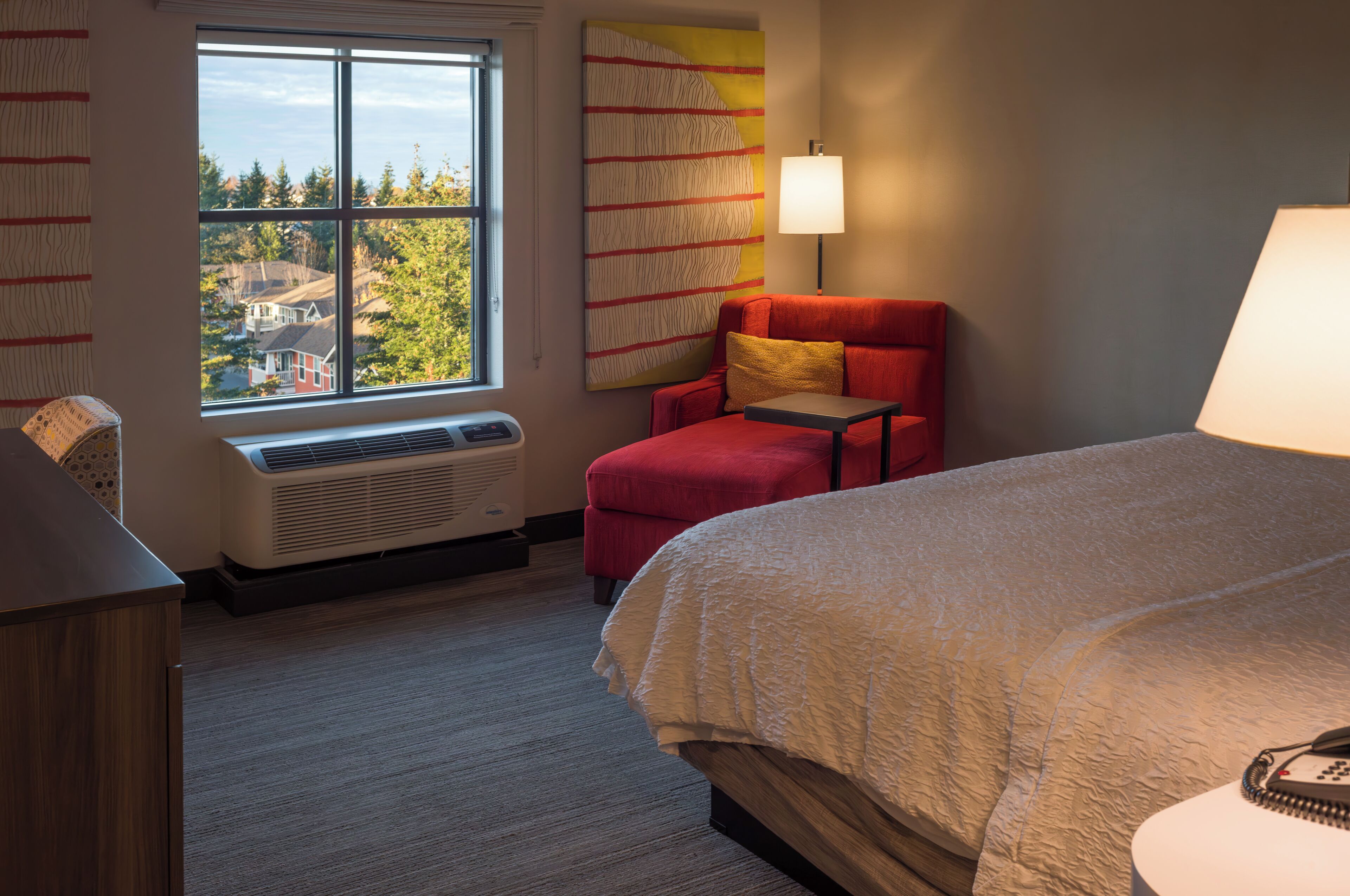 A hotel room with a large bed, a red armchair, a small table, a lamp, and a window with a view of houses and trees.