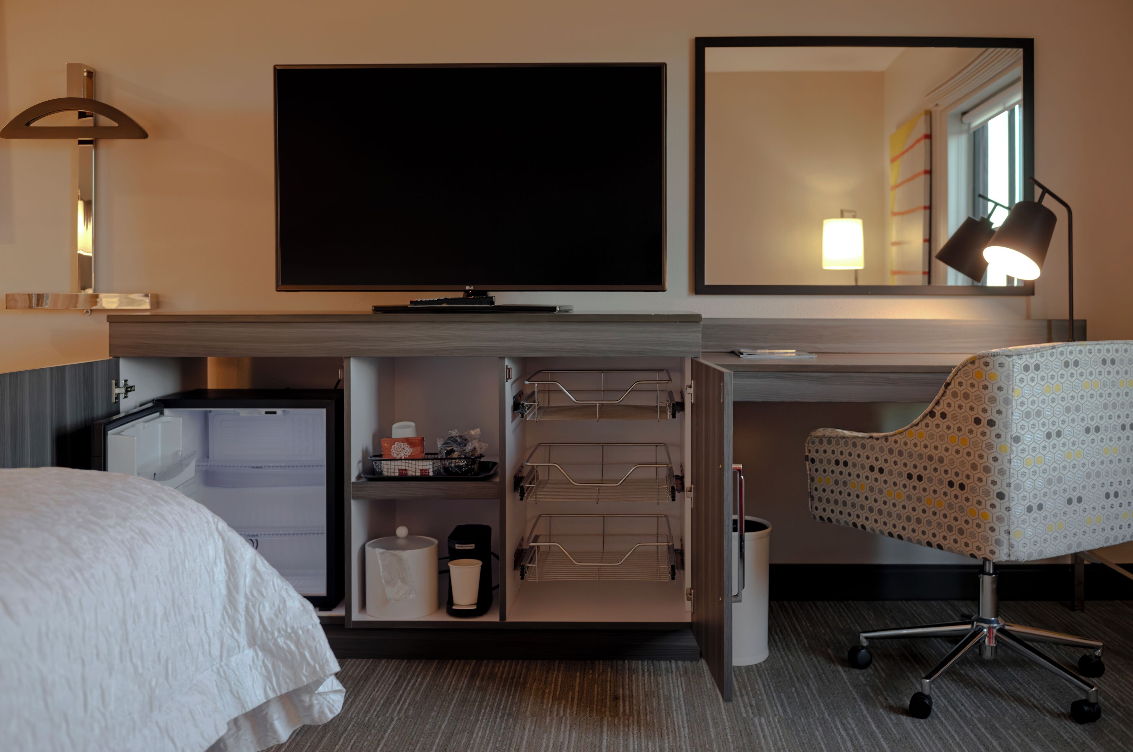 Habitación de hotel con cama, escritorio con silla, televisor, espejo, refrigerador y lámpara.
