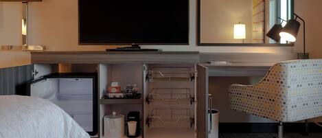 Habitación de hotel con cama, escritorio con silla, televisor, espejo, refrigerador y lámpara.