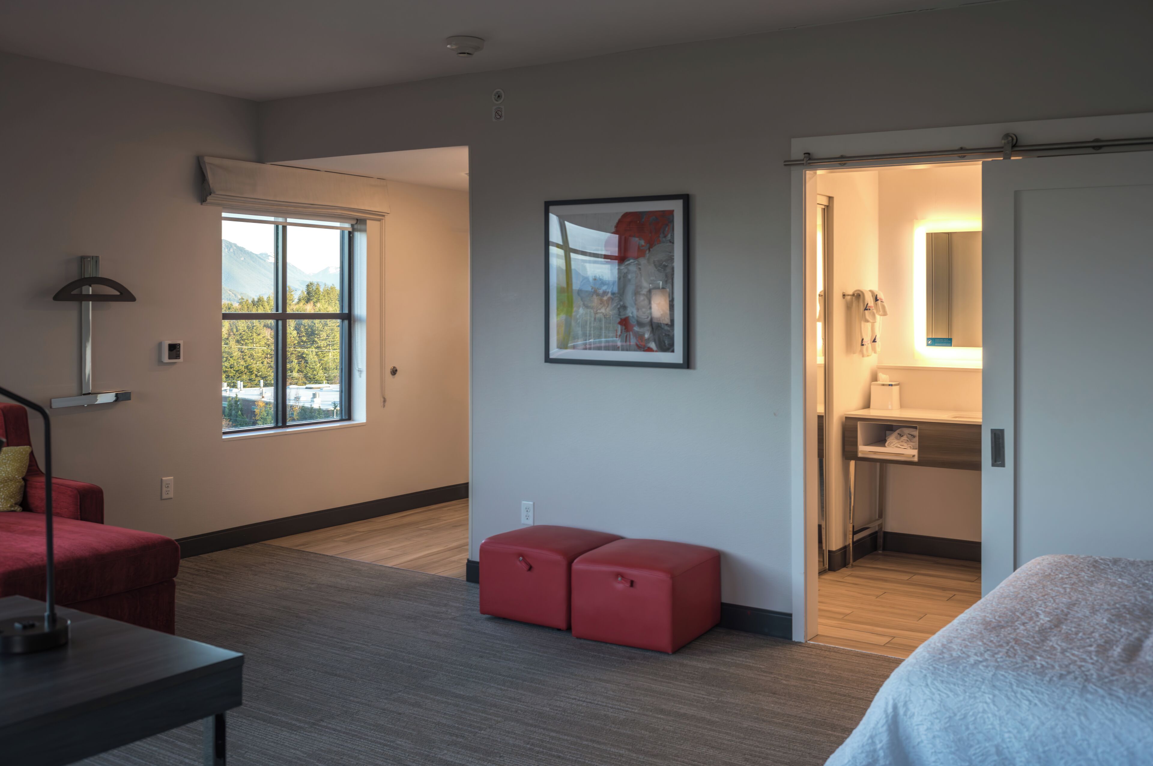 A modern hotel room with a large window, a red ottoman, a bed, a desk, and a bathroom with a mirror and towel rack.