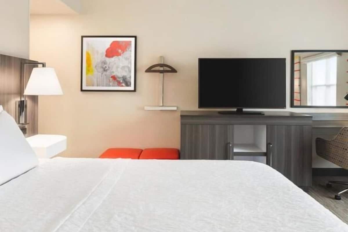 A hotel room with a large bed, a flat-screen TV, a desk with a chair, a lamp, and a framed abstract painting on the wall.