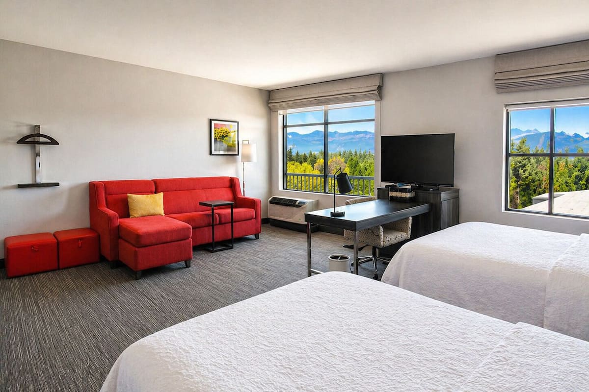 A hotel room with a red sofa, a desk with a TV, two beds, and a window with a mountain view.