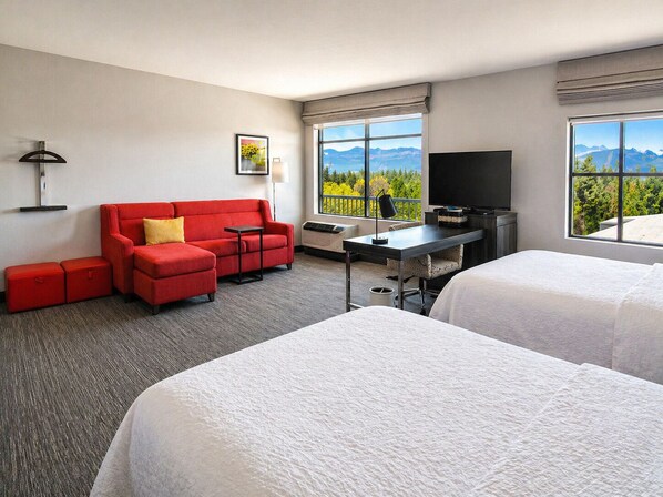 A hotel room with a red sofa, a desk with a TV, two beds, and a window with a mountain view.