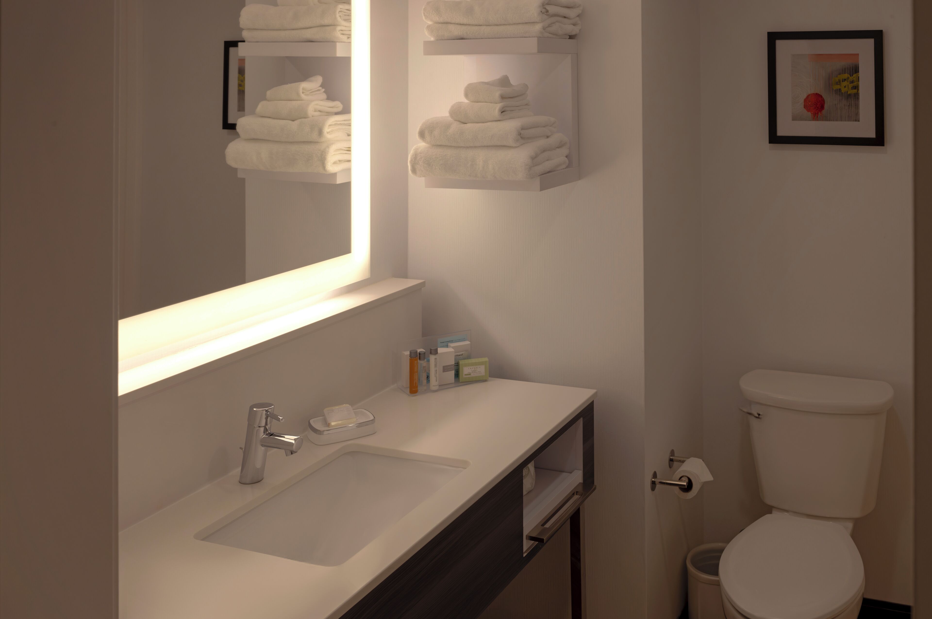 A modern bathroom with a white sink, mirror, and toilet. There are towels stacked on a shelf above the mirror and a framed picture on the wall.