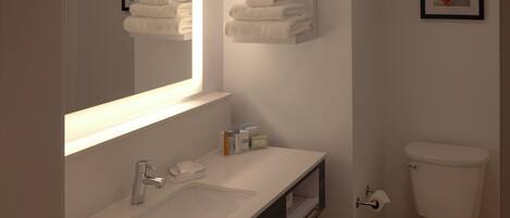 A modern bathroom with a white sink, mirror, and toilet. There are towels stacked on a shelf above the mirror and a framed picture on the wall.