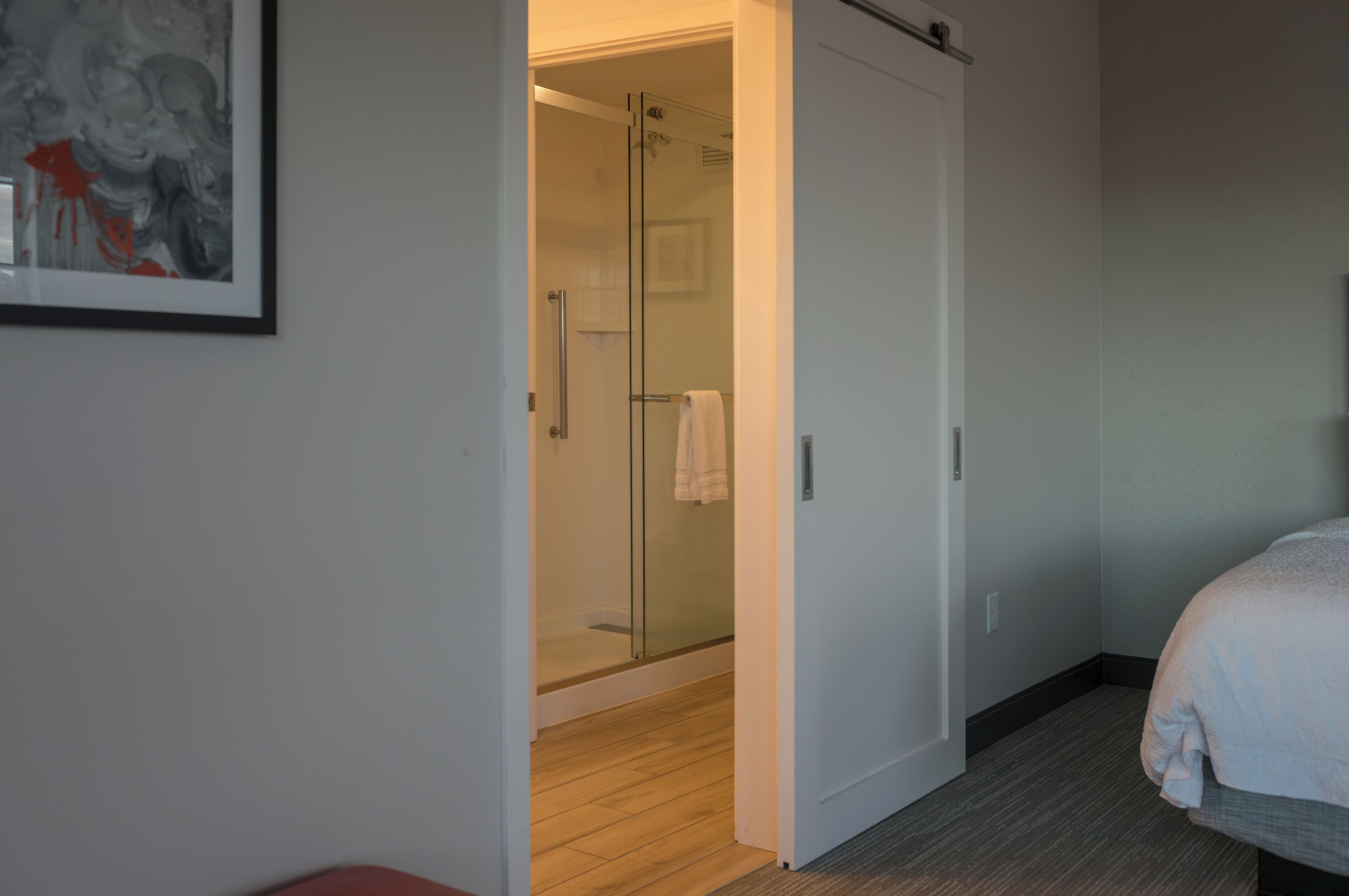 A hotel room with a glass shower door, a bed with a white cover, and a framed abstract painting on the wall.