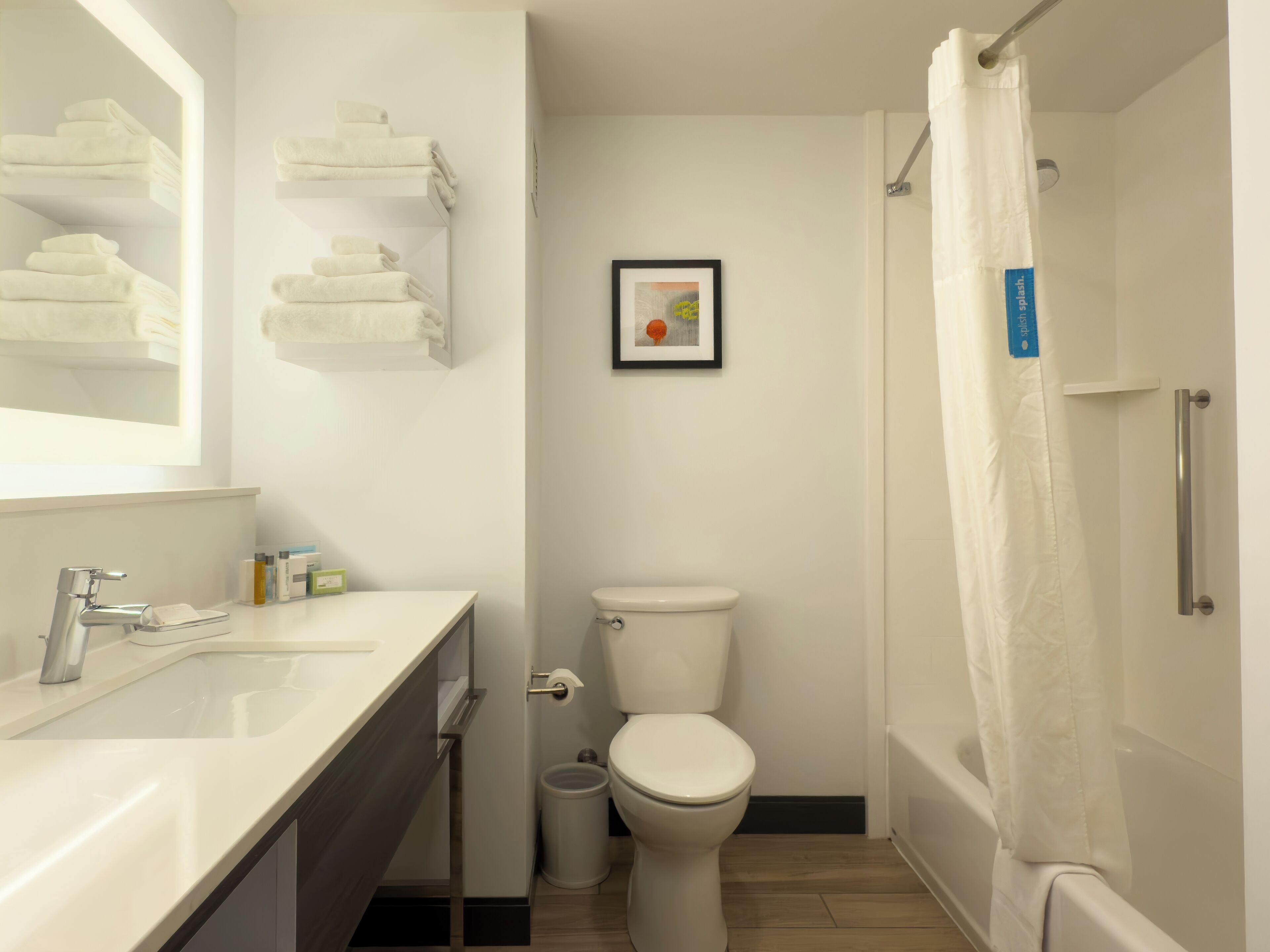 Ein modernes Badezimmer mit Waschbecken, Toilette und Dusche in Weiß. Handtücher liegen auf Regalen über dem Waschbecken gestapelt, und ein gerahmtes Bild hängt an der Wand.