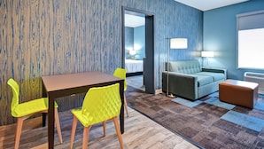 Habitación de hotel con pared de madera, una mesa de comedor con sillas amarillas, un sofá gris y un otoman marrón.