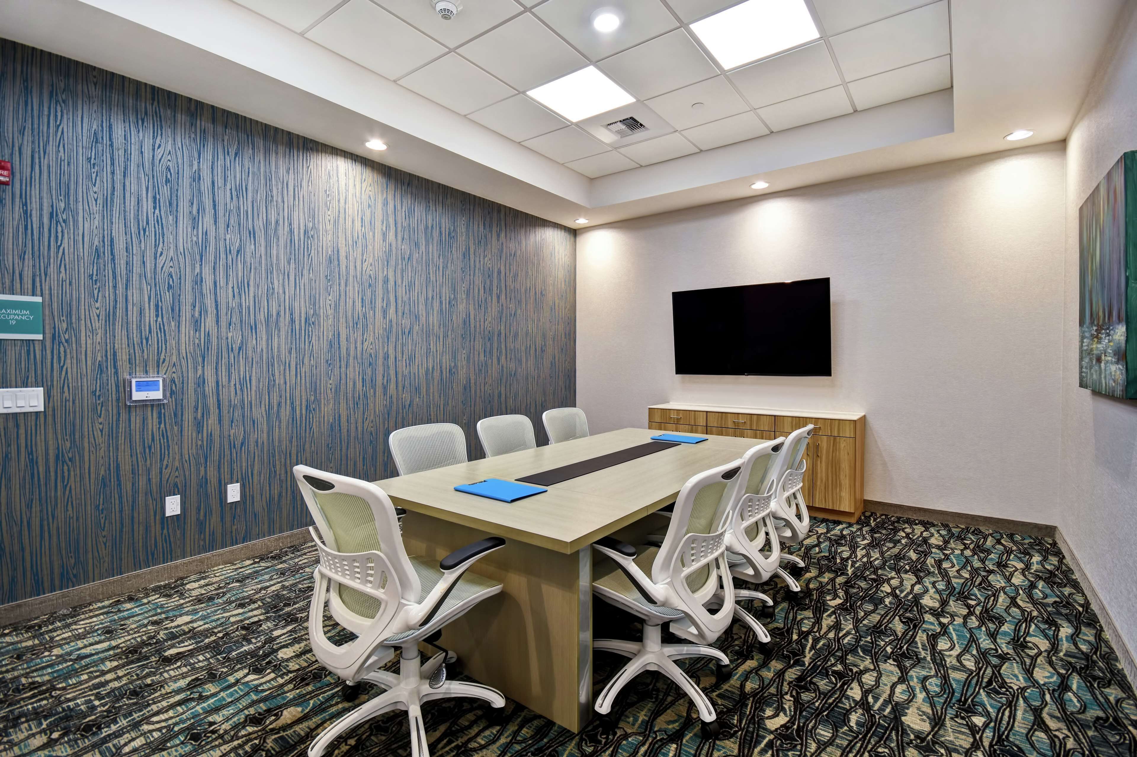 A conference room with a long table, white chairs, a flat-screen TV, and a patterned carpet.