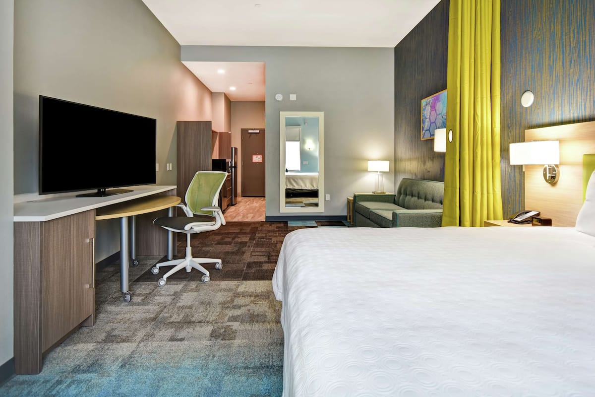 Habitación de hotel con cama, escritorio con televisor de pantalla plana, una silla, sofá, espejo y lámpara.
