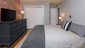 Een slaapkamer met een televisie op een dressoir, een bed met gestreepte lakens en een bakstenen muur.
